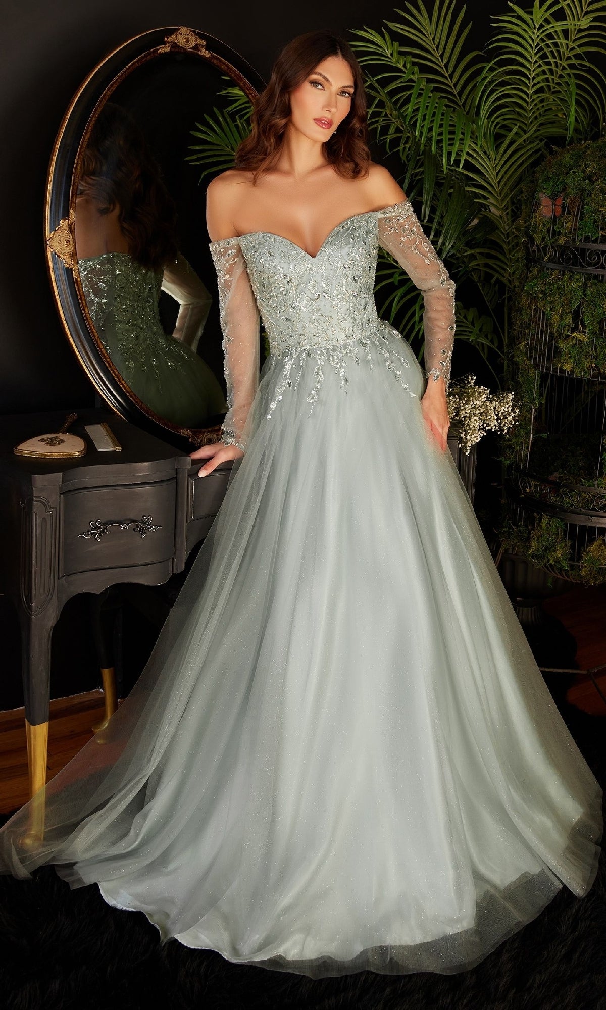 A woman stands indoors by a dark vanity and round mirror, wearing the Ladivine CD0172—a long sleeve, off-the-shoulder light blue prom dress with sheer sleeves and floral embellishments.